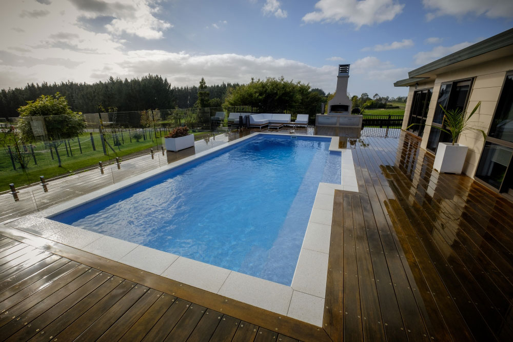 A luxurious backyard with a swimming pool and wooden deck beside a modern house. Outdoor fireplace, seating area, and lush green surroundings visible.