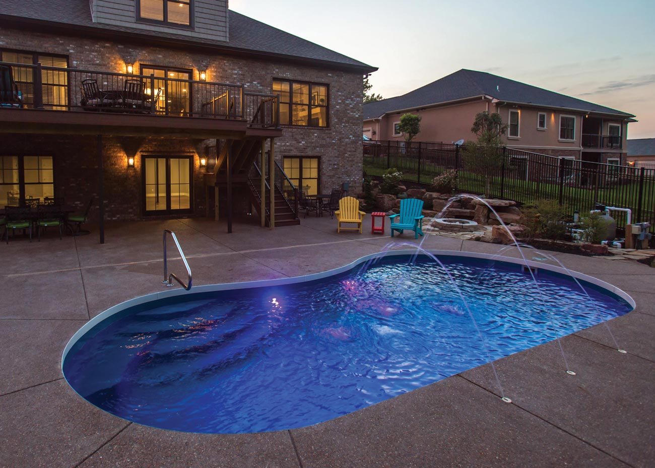 A kidney-shaped swimming pool glows at dusk outside a large multi-story house with lights on, patio furniture around, and a neighboring house visible.