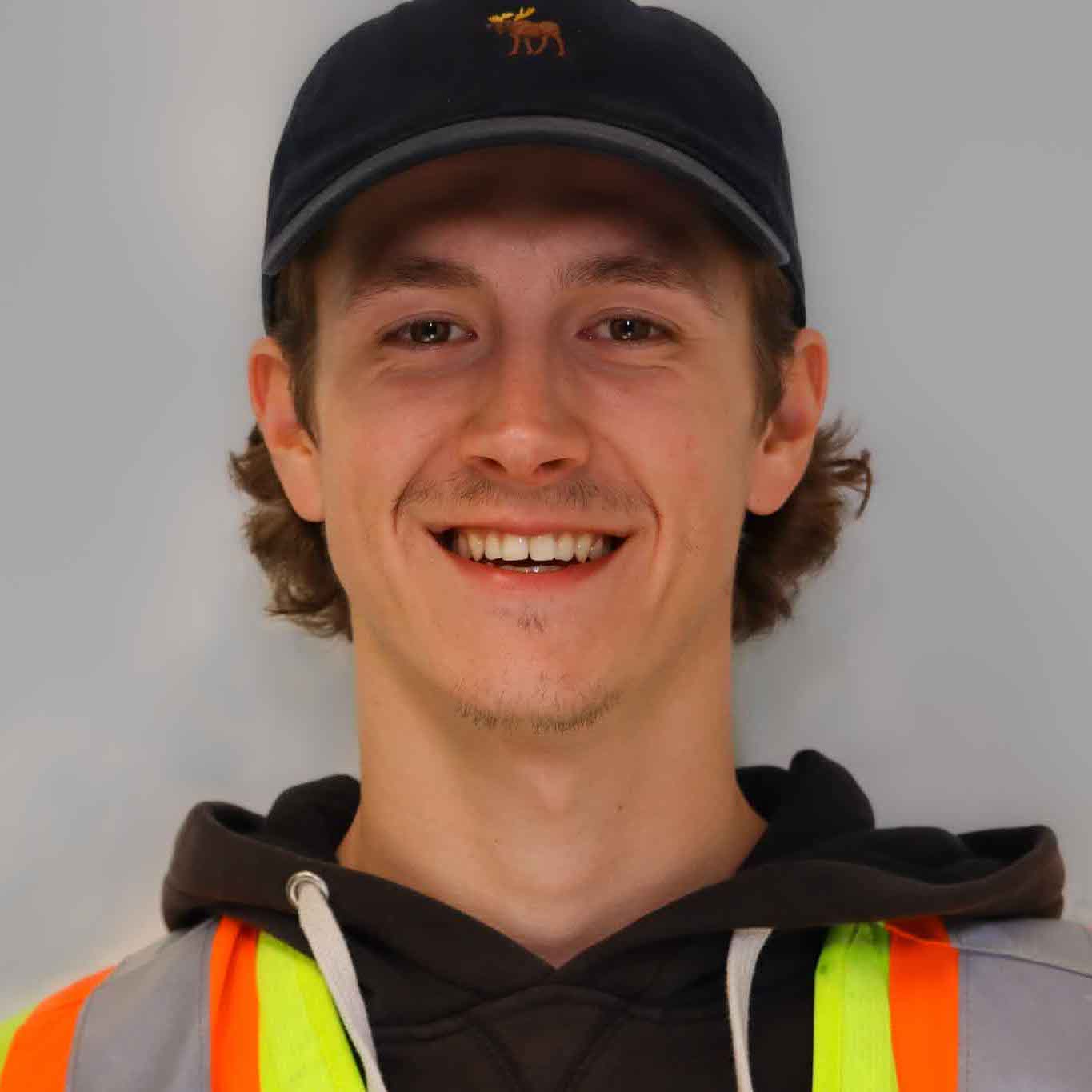 A person wearing a cap and a high-visibility safety vest smiles in front of a gray background. The individual appears cheerful and casual.