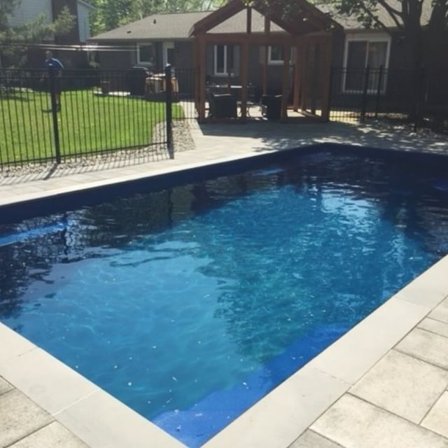 A rectangular swimming pool with blue water is surrounded by a patio and a fence, with a gazebo structure and greenery in the background.