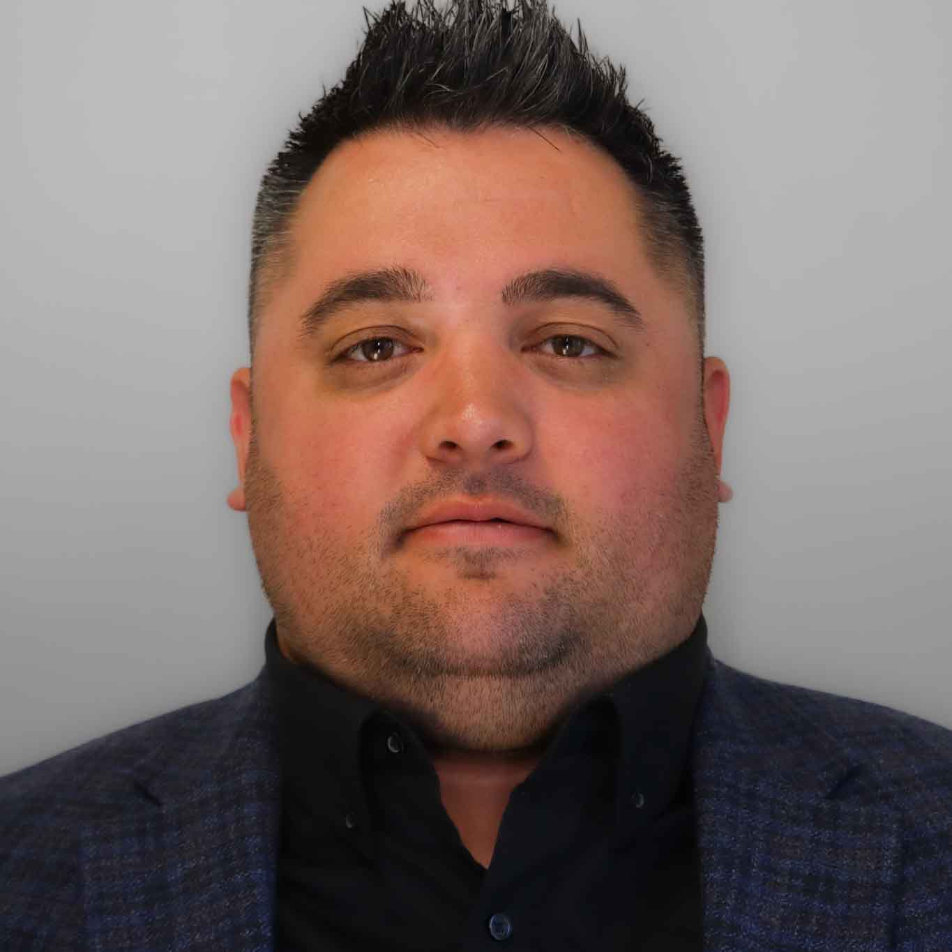 The image shows a person with spiked hair, a light beard, wearing a black shirt under a patterned blazer, and posing against a grey backdrop.