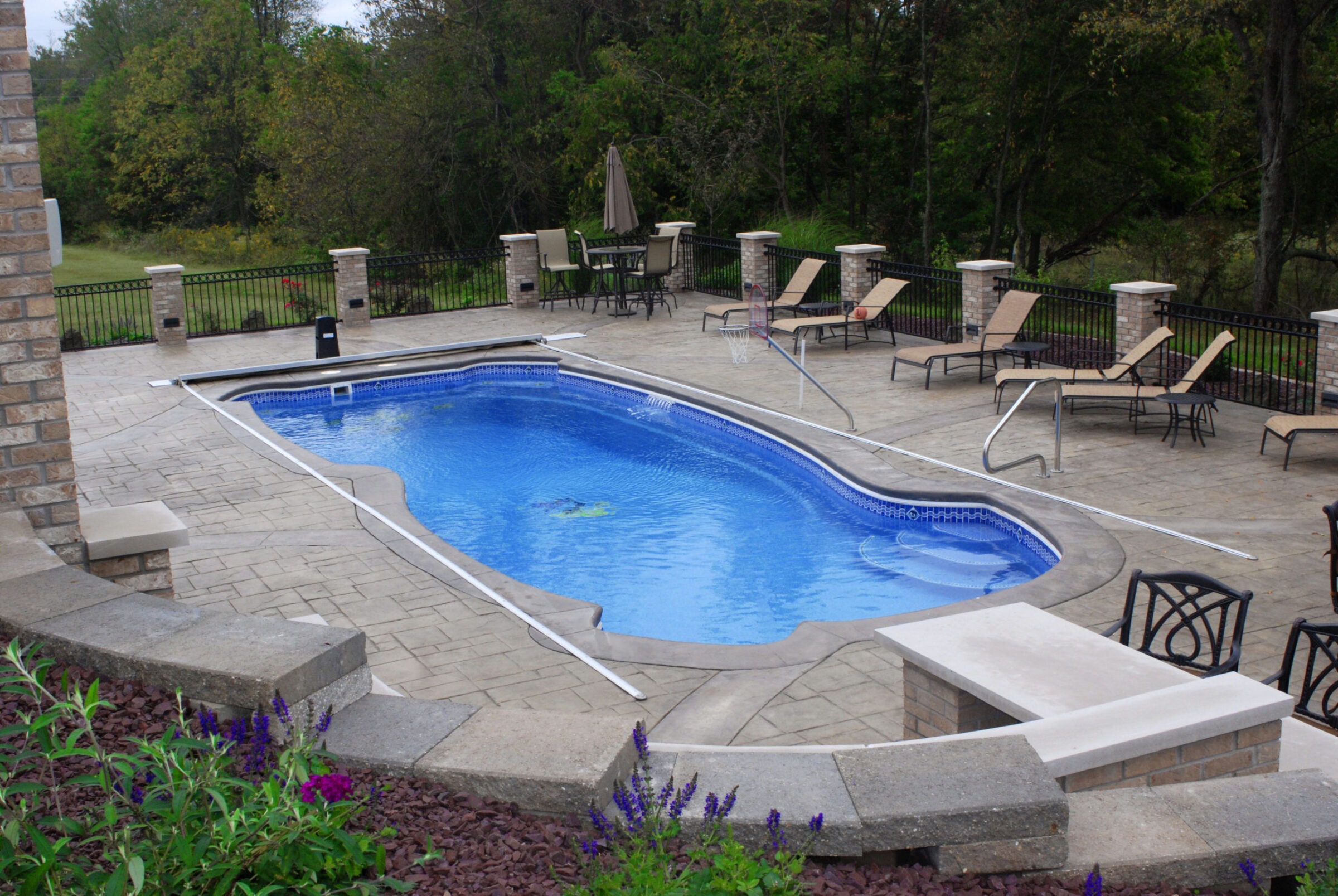 An outdoor swimming pool with blue water, surrounded by patio furniture, umbrellas, and landscaping, enclosed by a wrought-iron fence amidst natural greenery.