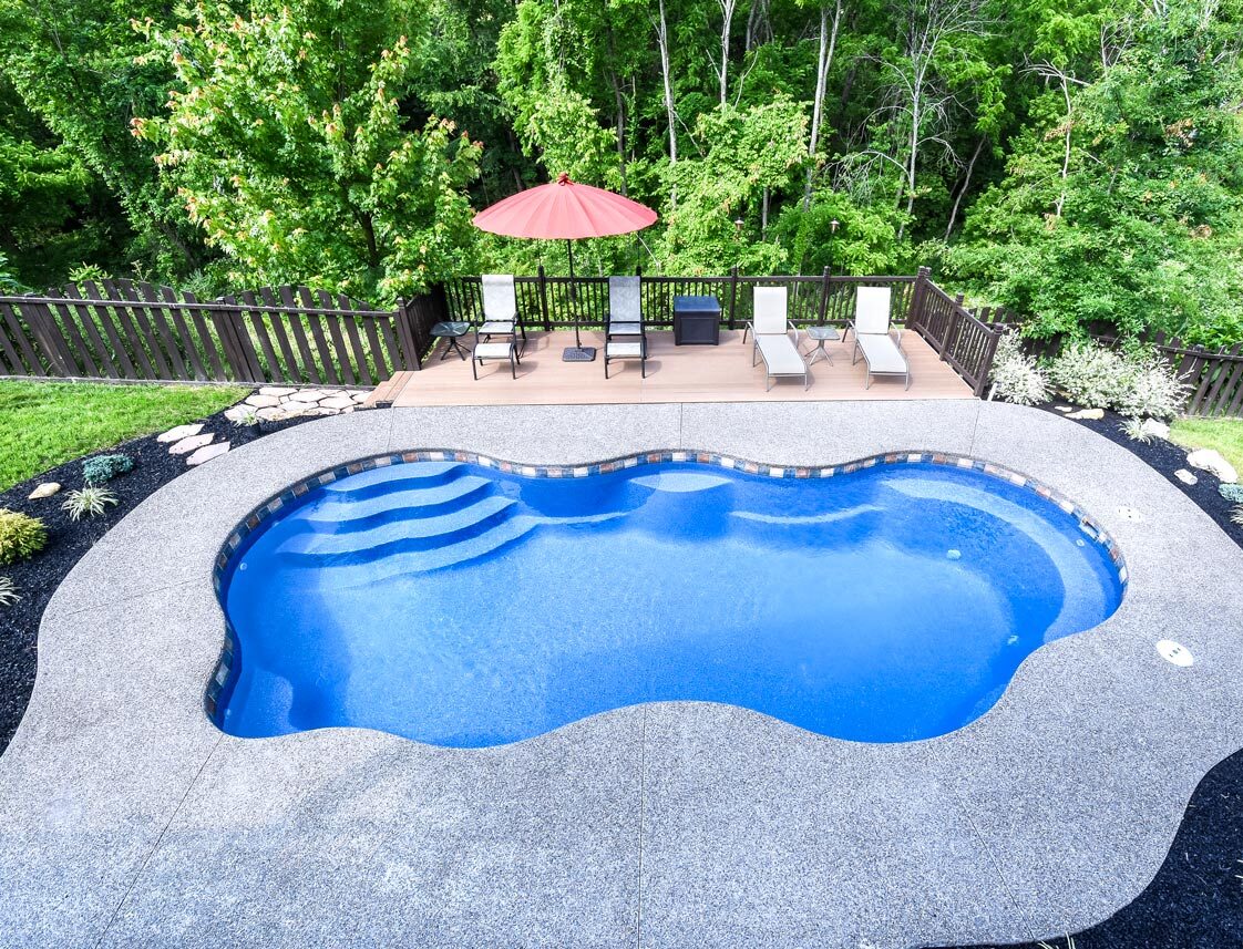 Kiddie blue swimming pool with an irregular shape, surrounded by a deck and lounge chairs under a red umbrella, set against lush green trees.