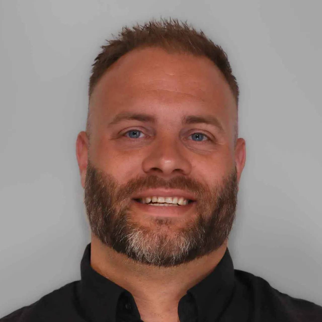 A person with short hair, a beard, and blue eyes is smiling. They're wearing a black shirt and have a neutral background.