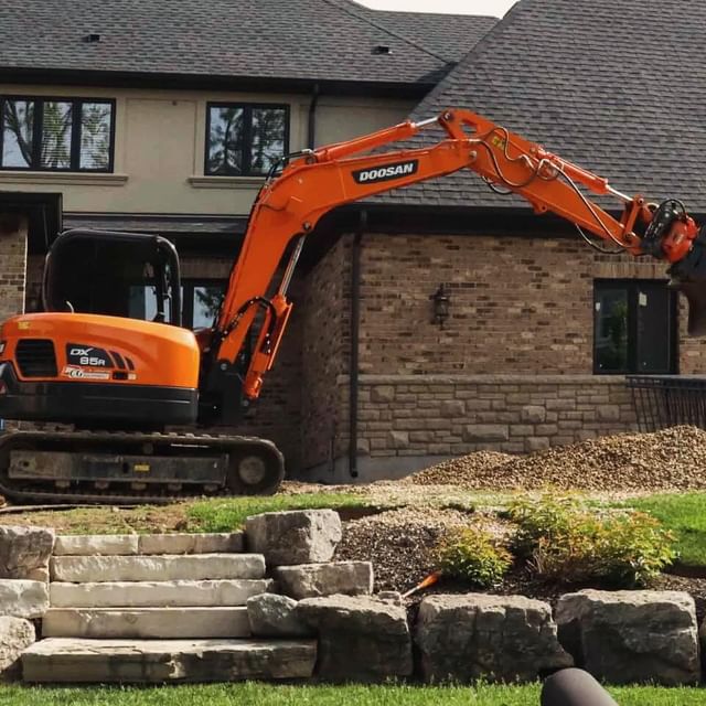 An orange Doosan excavator parked by a residential house with stone steps and landscaping, indicating construction or renovation work nearby.