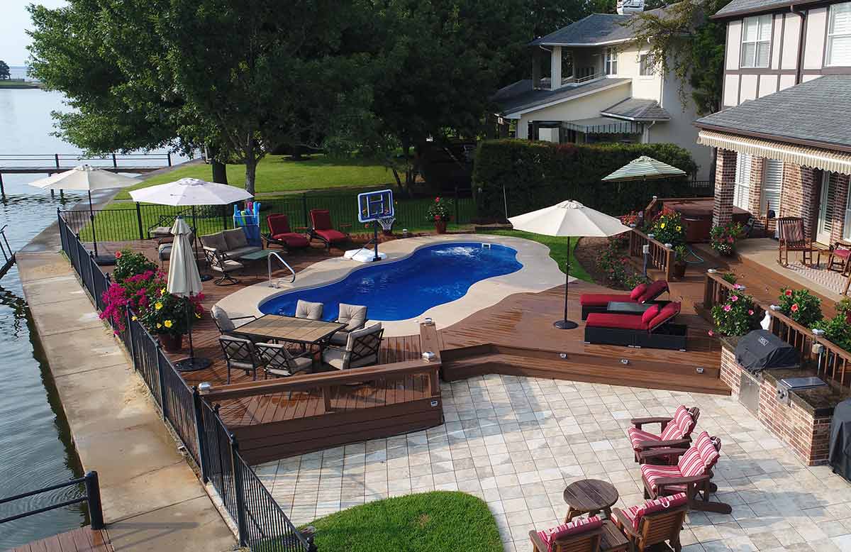 A luxurious backyard featuring an in-ground swimming pool, patio furniture, umbrellas, a wooden deck, and a green lawn overlooking a body of water.