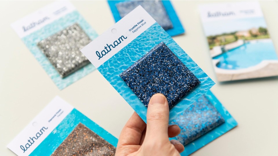 A person's hand is holding a sample of blue pool liner material among several samples with different textures and colors on a light background.