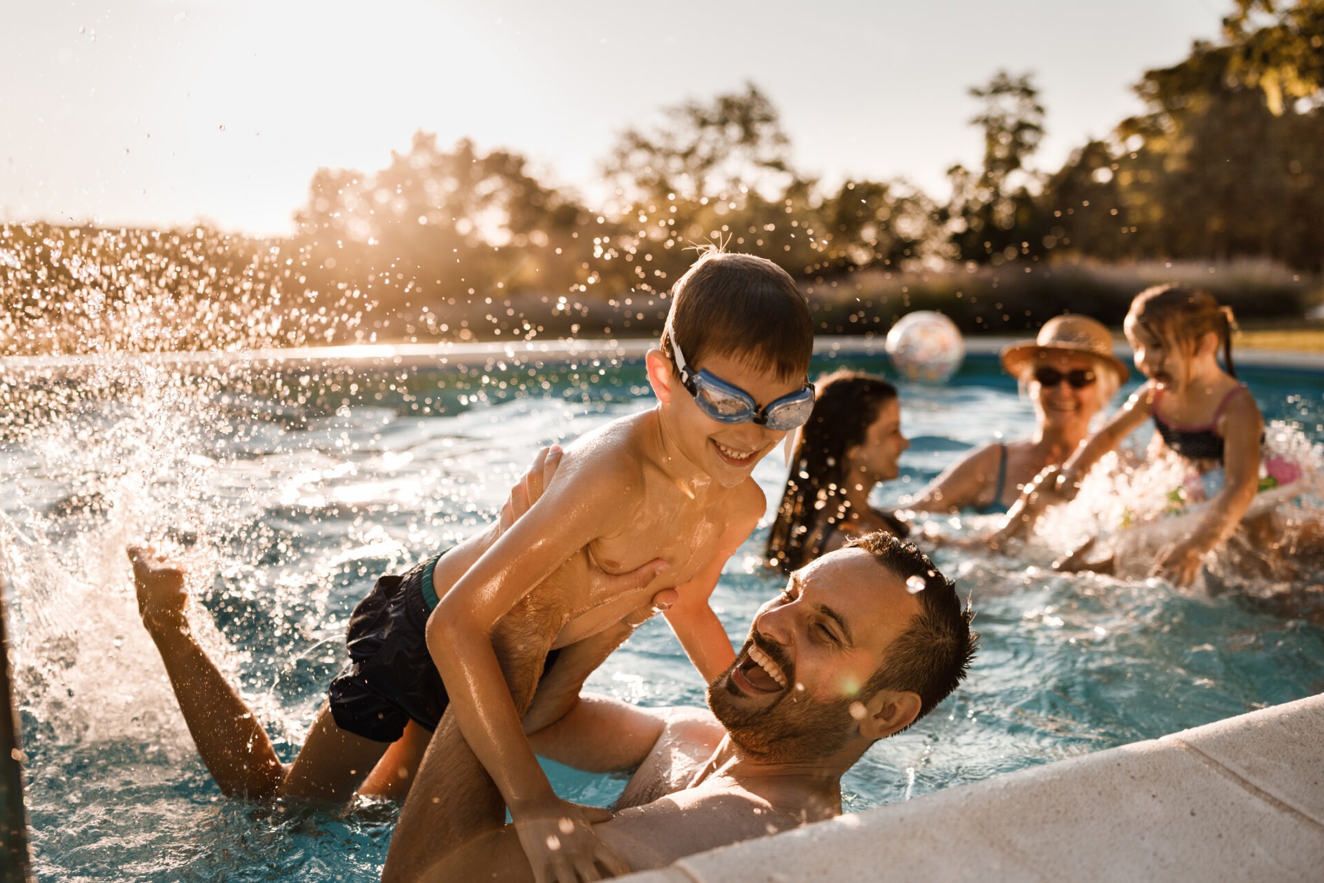 A joyful scene in a sunlit pool with a person lifting a child on their shoulders while others splash water, capturing moments of laughter and play.