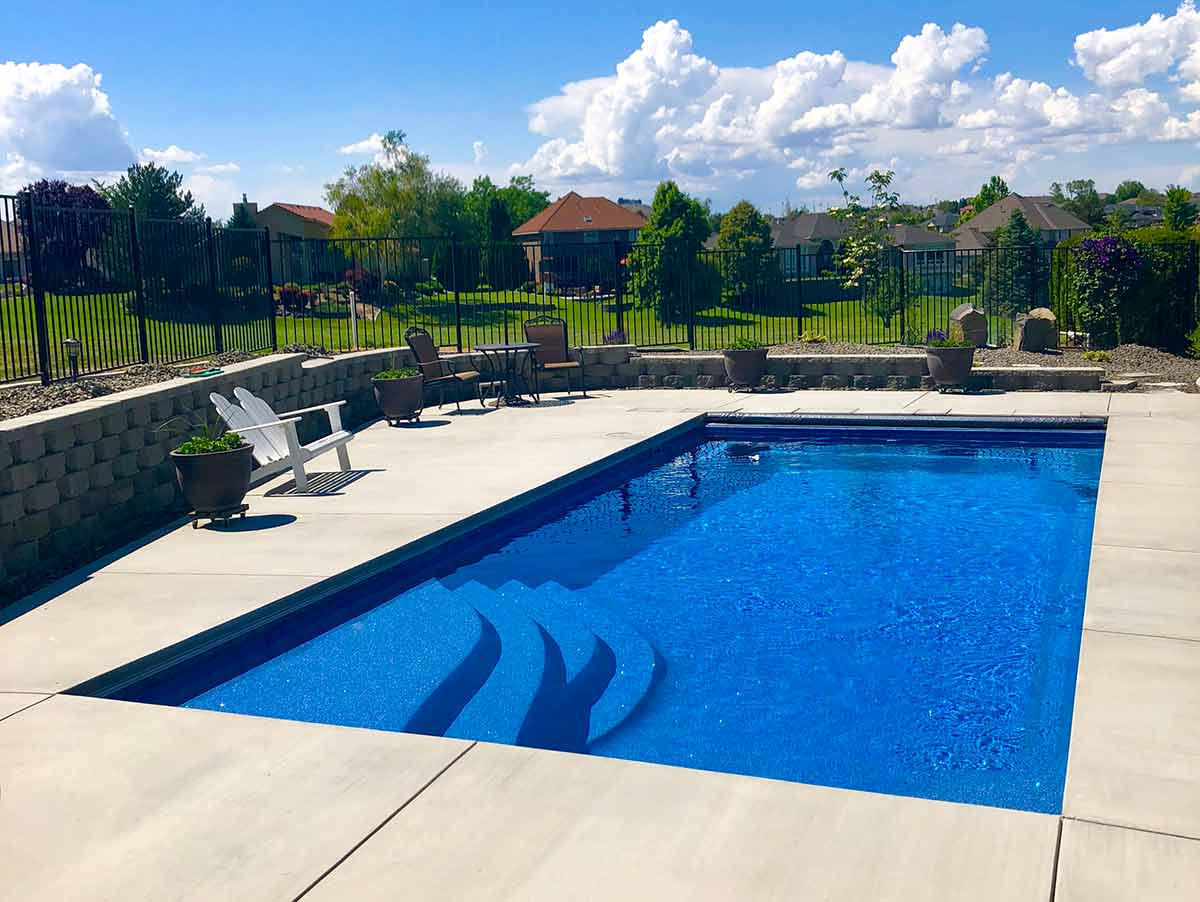 An outdoor swimming pool with clear blue water is surrounded by patio chairs, potted plants, and a neatly landscaped lawn with a fence.