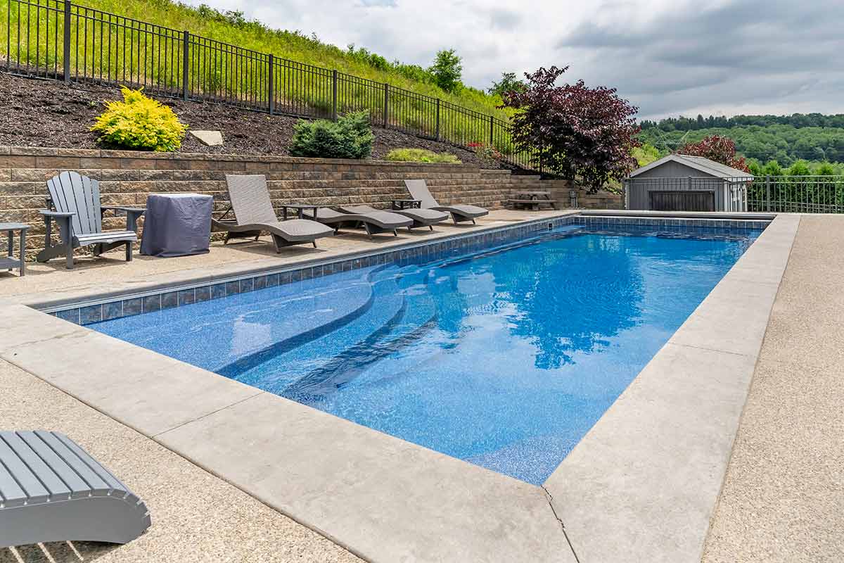 An inground swimming pool with clear blue water, surrounded by lounge chairs and a lush hillside with a fence at a residential property.