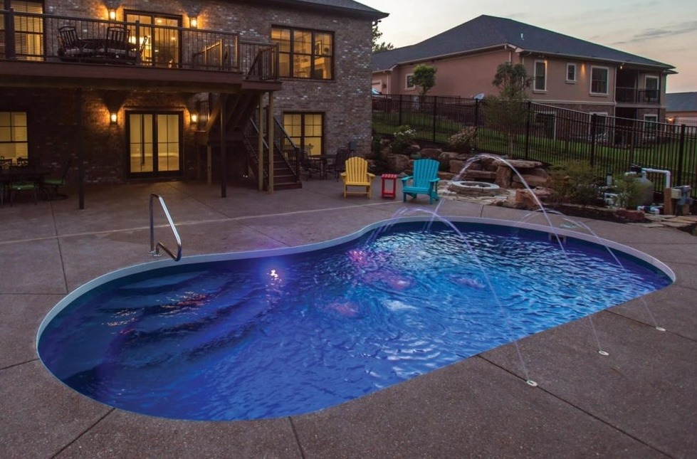 This image shows a kidney-shaped swimming pool with clear blue water, surrounded by a patio area and a multi-story brick house with balconies.