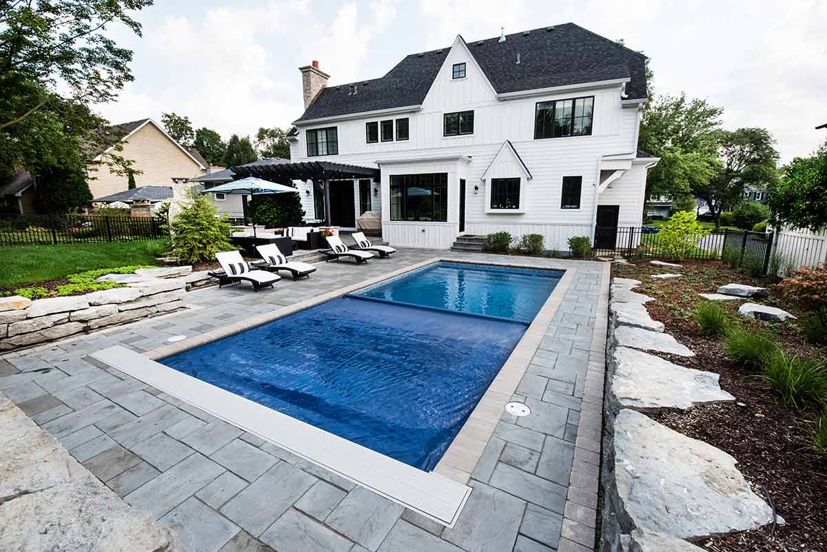 A serene backyard with a rectangular swimming pool, lounge chairs, landscaped garden, a two-story house with white siding, and a covered patio area.