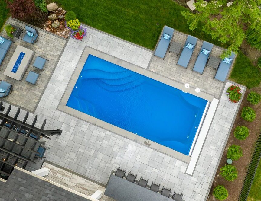 Aerial view of a backyard with a rectangular pool, surrounded by patio furniture and neatly landscaped garden areas with green grass and plants.