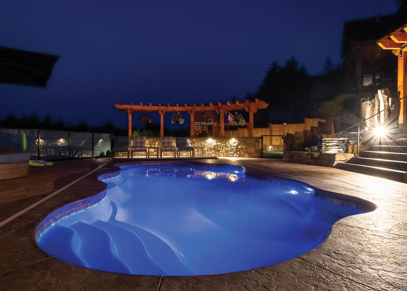 An illuminated outdoor swimming pool at dusk with adjacent lounge chairs, stone accents, a pergola, and a wooded backdrop under a twilight sky.