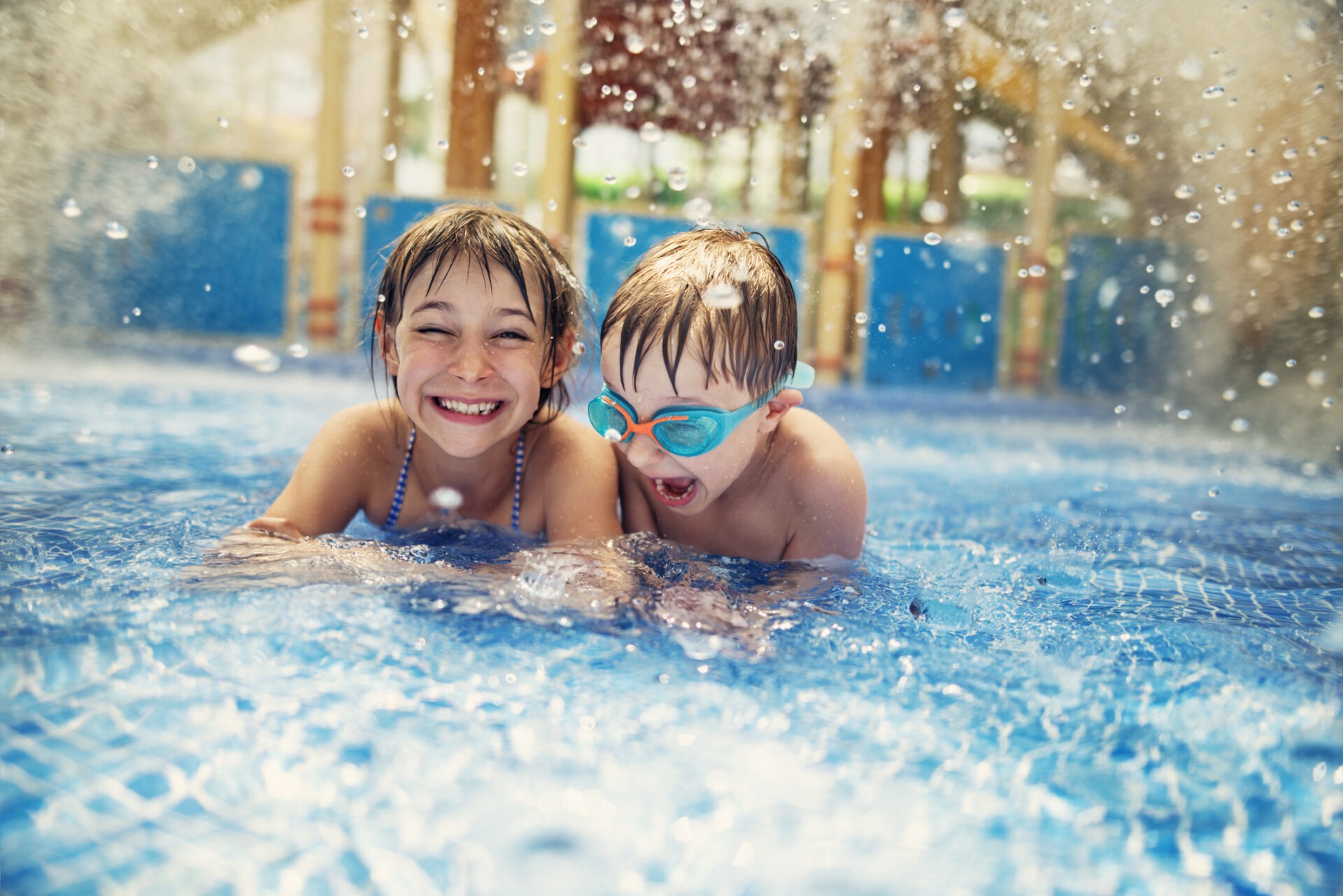 Two joyful children are splashing in a sunlit pool, one wearing goggles; the water sparkles with droplets flying through the air.