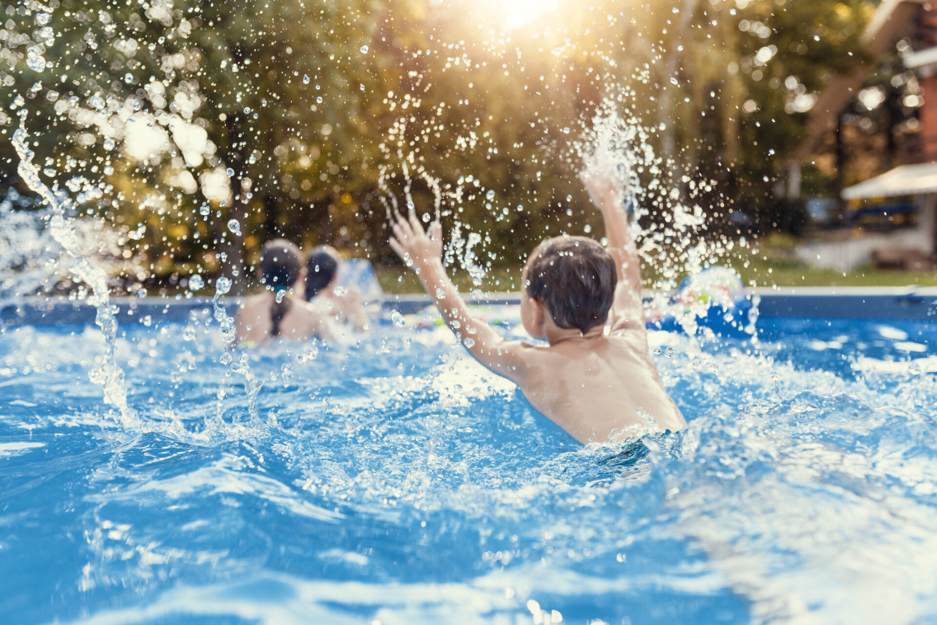 A child splashes in a sunlit outdoor pool with two people in the background. Water droplets are suspended in the air, capturing the playful moment.