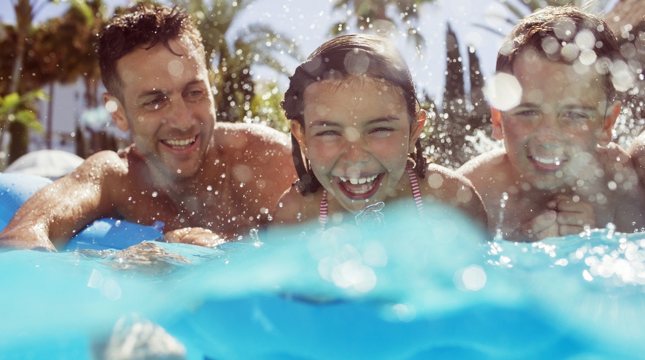 A person and two children are smiling in a sunlit swimming pool, surrounded by splashing water droplets, with palm trees in the background.