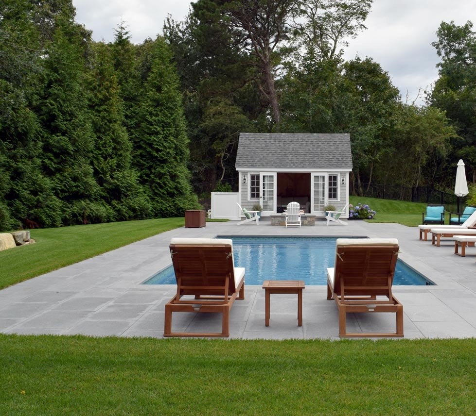 An idyllic backyard setting with a swimming pool, two lounge chairs, a small pool house, green lawn, and a dense backdrop of tall trees.